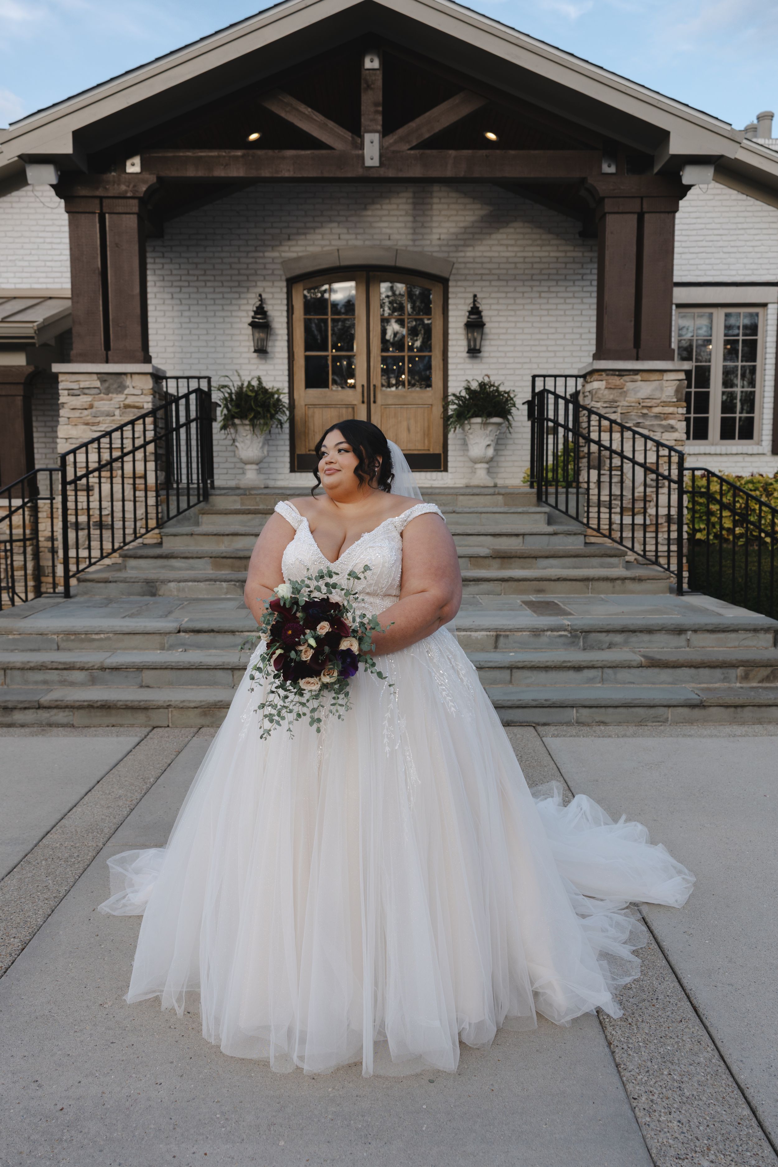 A beautiful wedding dress with a full tulle skirt and off-shoulder neckline photographed on stone steps outside a venue.