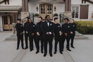 A group of groomsmen in black suits pose together outside a building entrance.