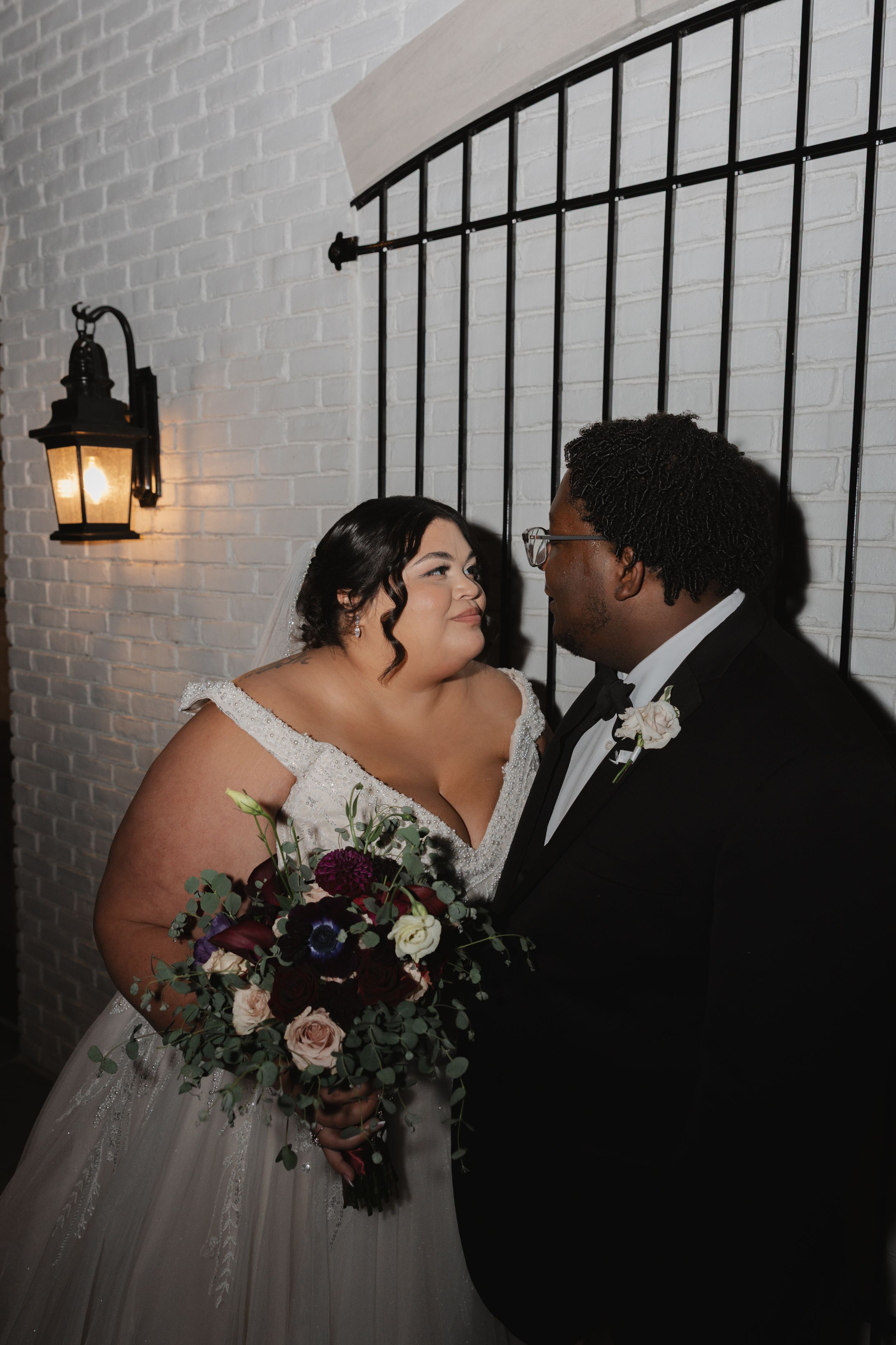 A romantic moment between newlyweds against a white brick wall with a vintage lantern glowing nearby.