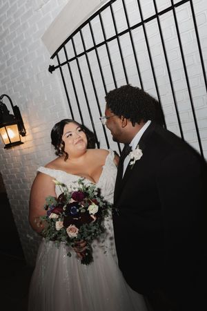 A romantic wedding moment between a couple in front of a white brick wall with decorative lighting.