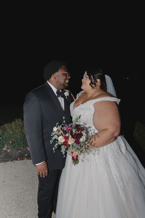 A romantic nighttime wedding portrait showing a couple embracing in formal attire with colorful floral bouquet.