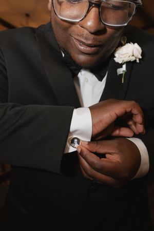 A groom in a black tuxedo and glasses adjusts his cufflink while wearing a white boutonniere.