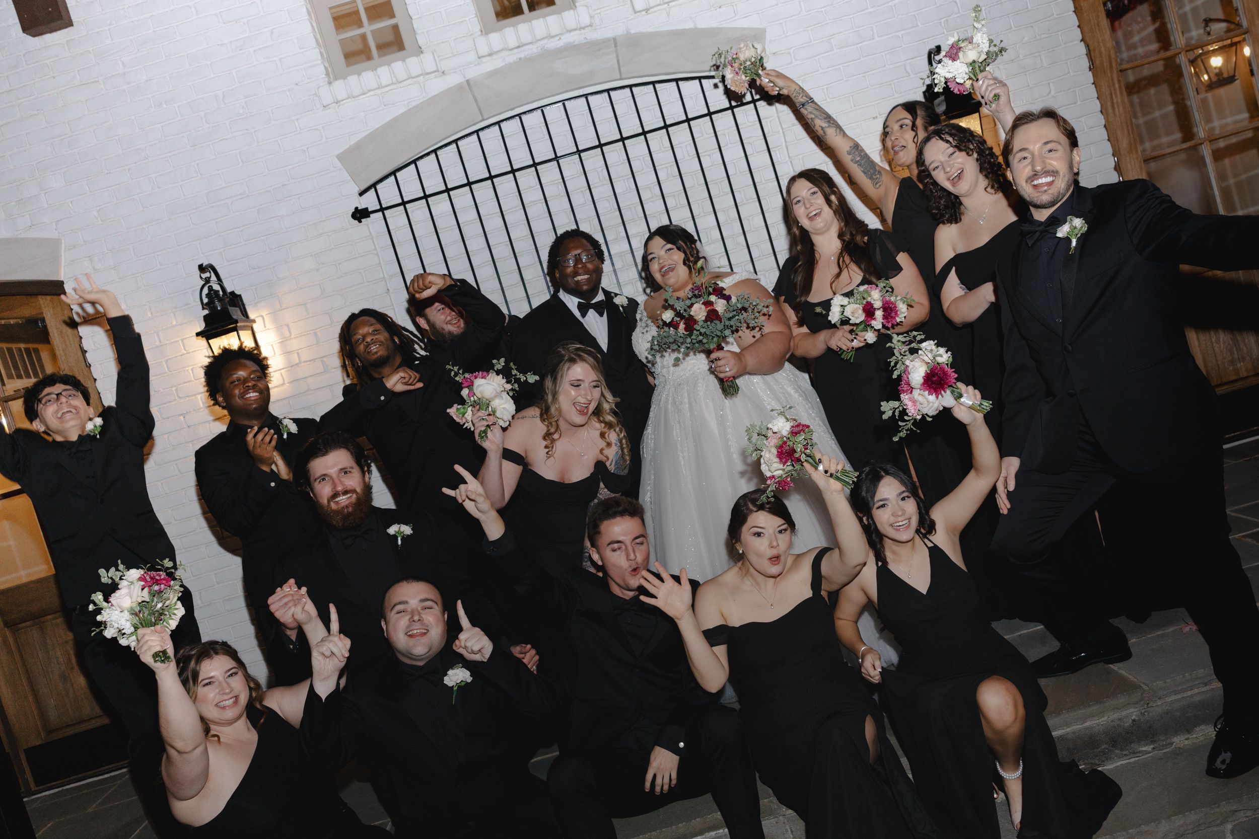 Wedding party celebrating with raised arms and bouquets outside a venue with an iron gate.