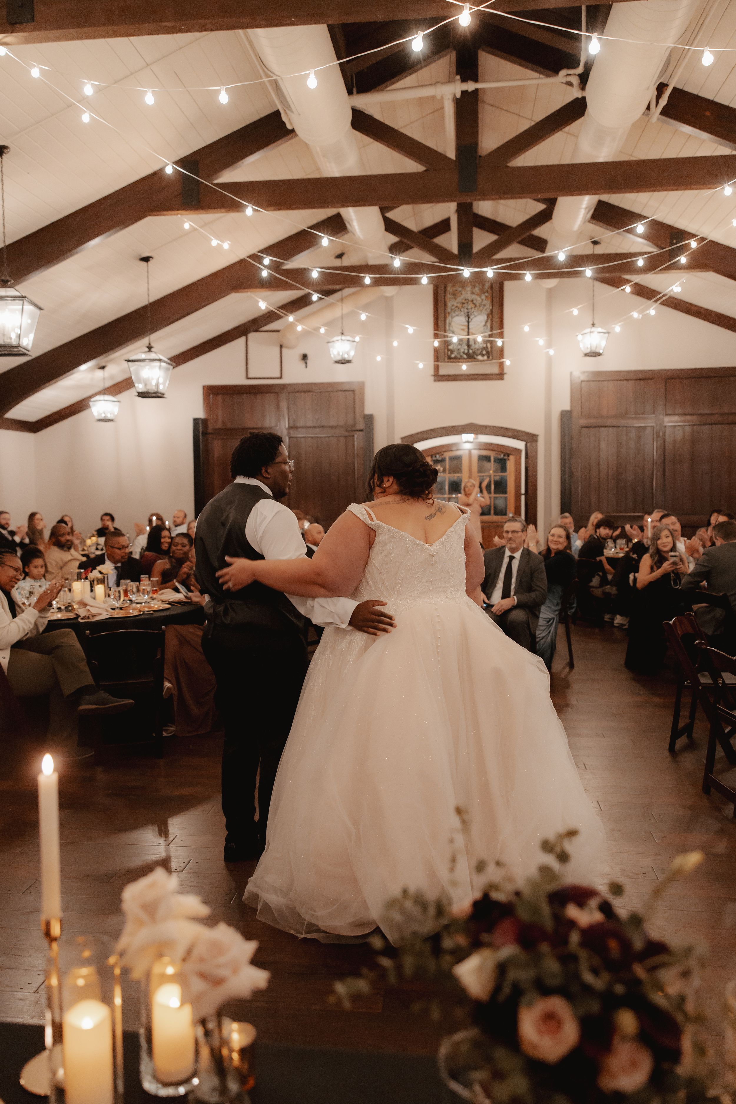 A romantic first dance in an elegant reception hall with exposed beams and string lights.