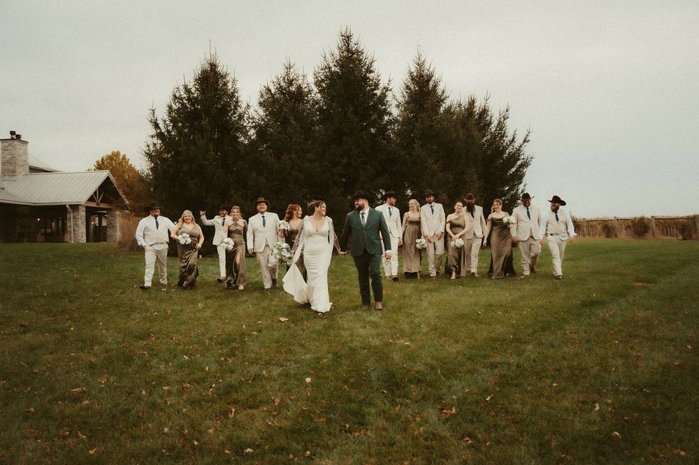 A group of people in white clothing gather outdoors for a panoramic photo in a grassy field.