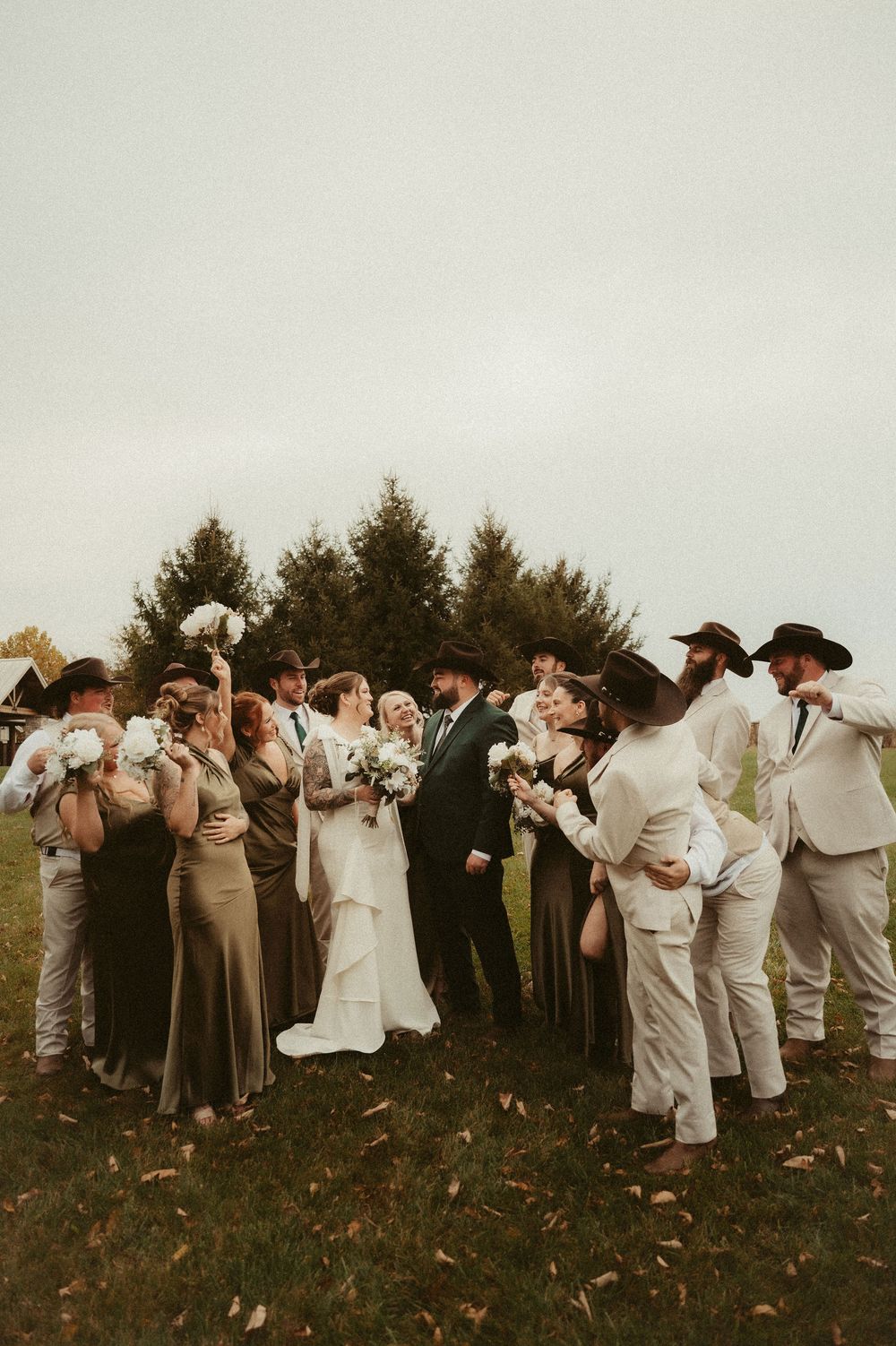 Wedding party members in neutral colors stand in line on a grassy hill while taking photos at sunset.