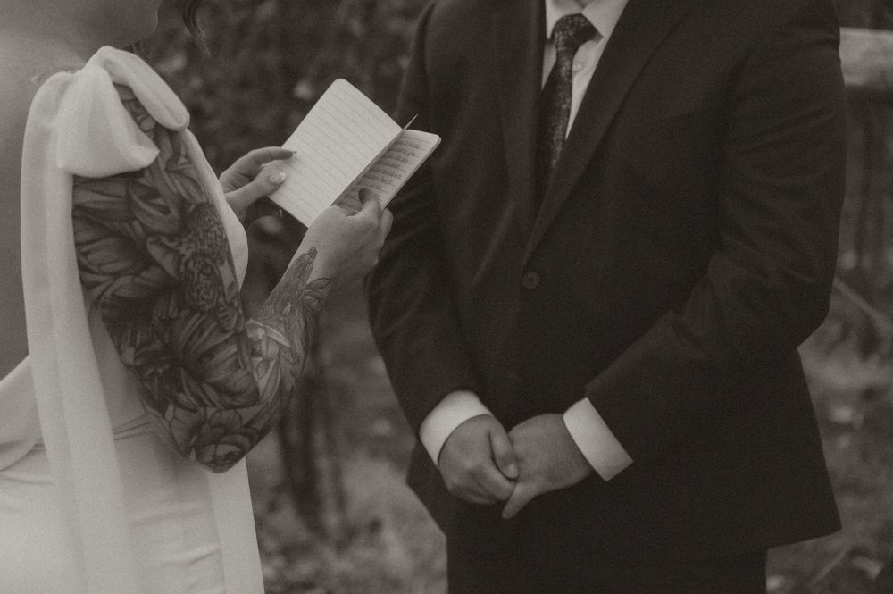 Black and white wedding photo showing guests reading from paper programs during an outdoor ceremony.