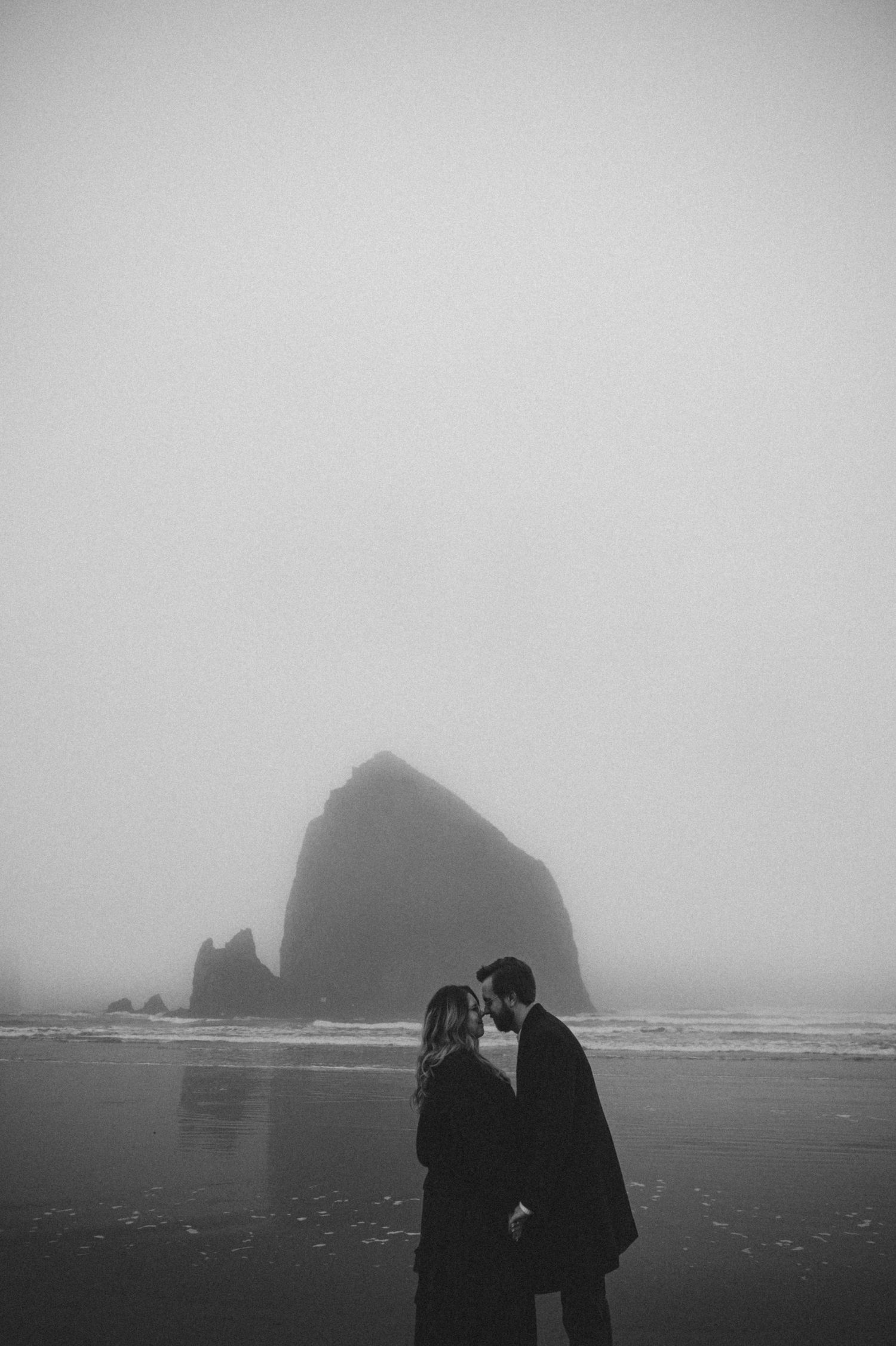 Silhouettes of two people embracing on a misty beach with dramatic rock formations in the background.