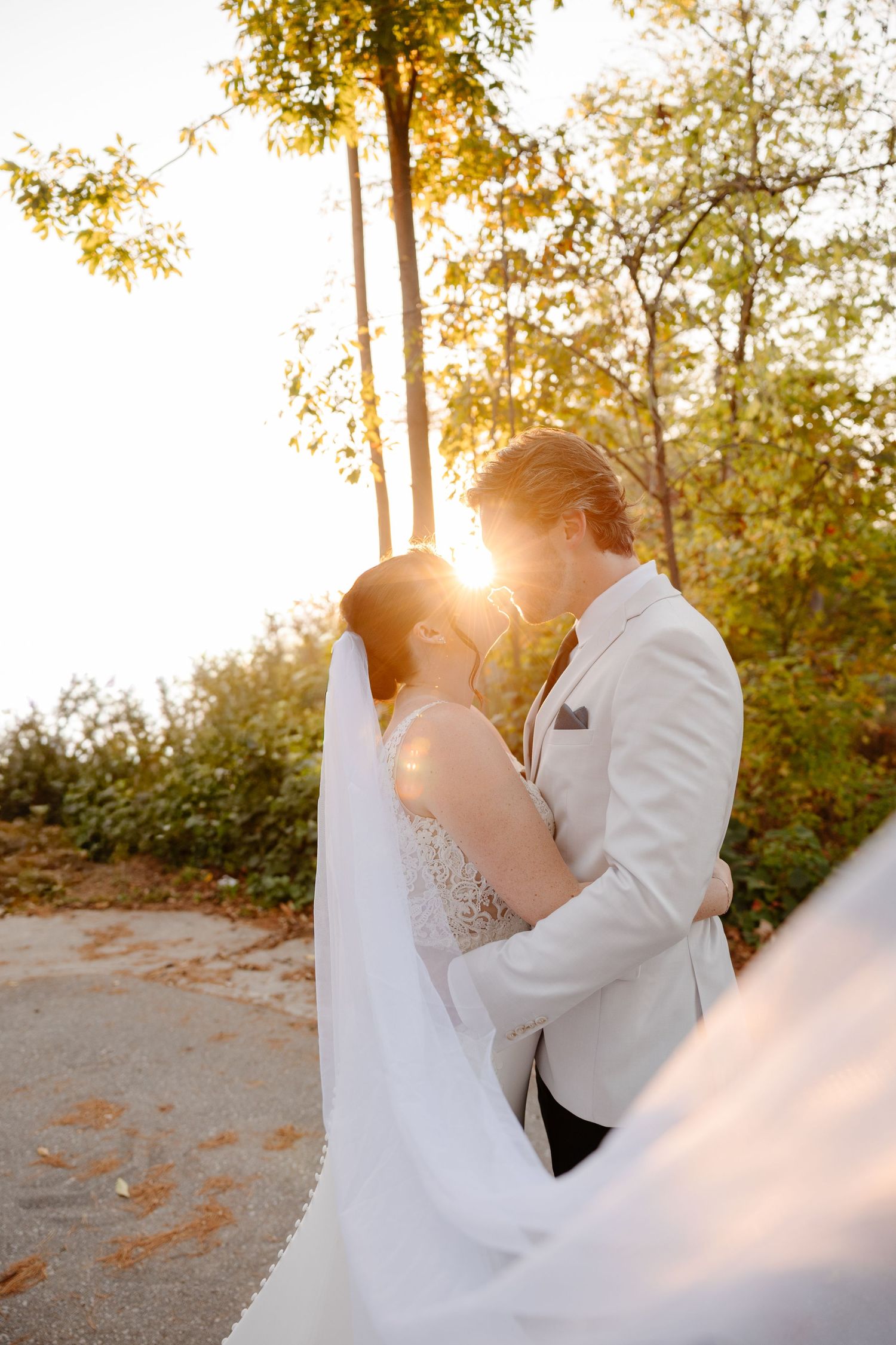 A romantic wedding moment captured at golden hour with sunlight streaming through autumn trees as a couple embraces.