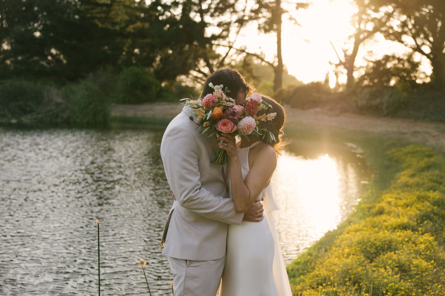A couple in wedding attire embraces during golden hour sunset by a serene lake creating a romantic silhouette.
