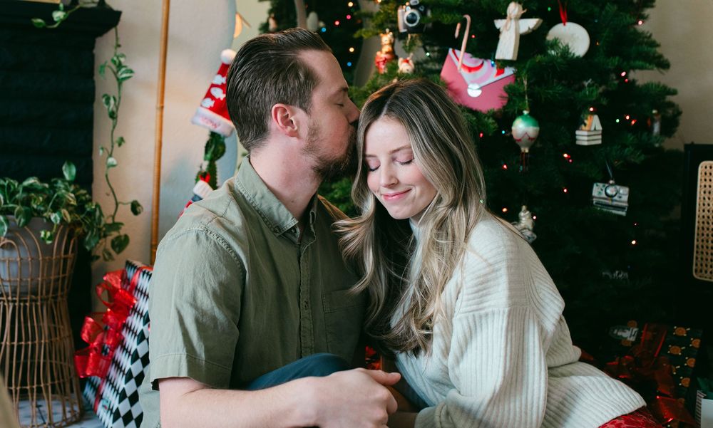 A couple shares a romantic moment in front of a decorated Christmas tree in a cozy living room setting.