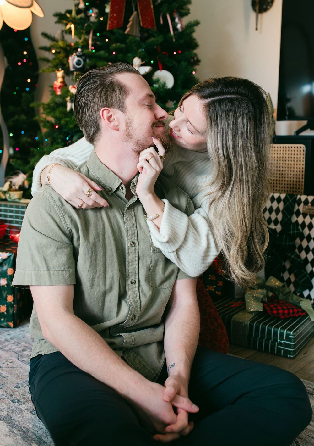 A romantic holiday moment shared between a couple in front of a decorated Christmas tree with wrapped presents nearby.