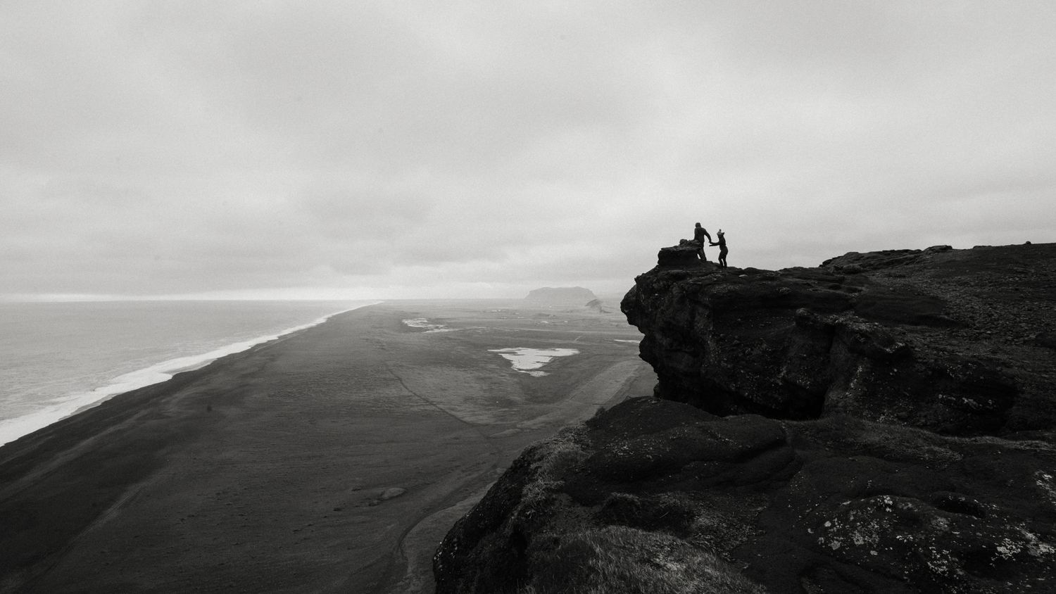 Two tiny figures stand on a dramatic black volcanic cliff overlooking a vast black sand beach in black and white.