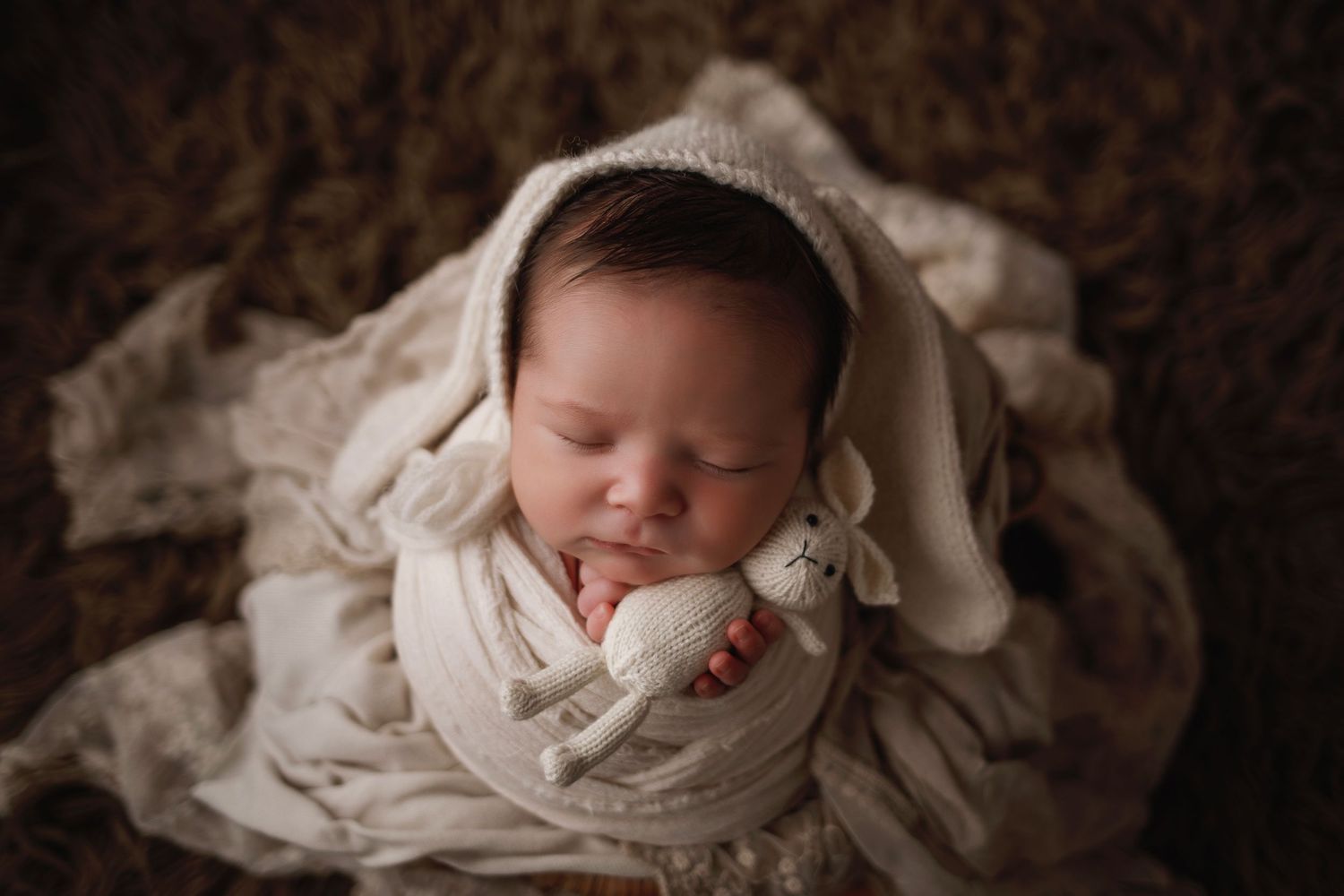 A sleeping newborn baby wearing cream-colored swaddle wrap lies peacefully in a cozy brown setting.
