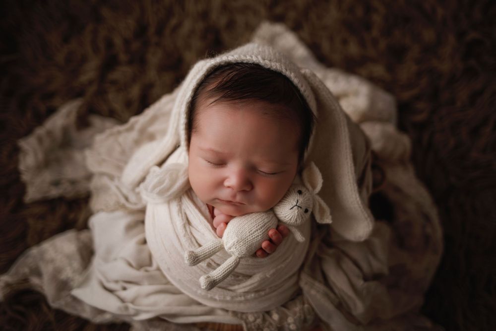 A sleeping newborn baby wearing cream-colored swaddle wrap lies peacefully in a cozy brown setting.