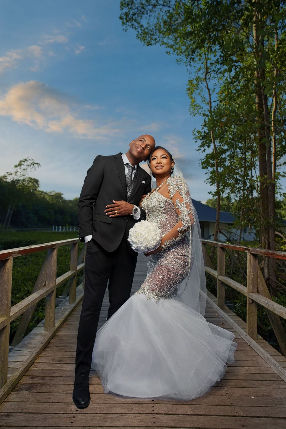 Bride and groom pose on wooden bridge, she in beaded mermaid gown with veil, he in black suit, at sunset.