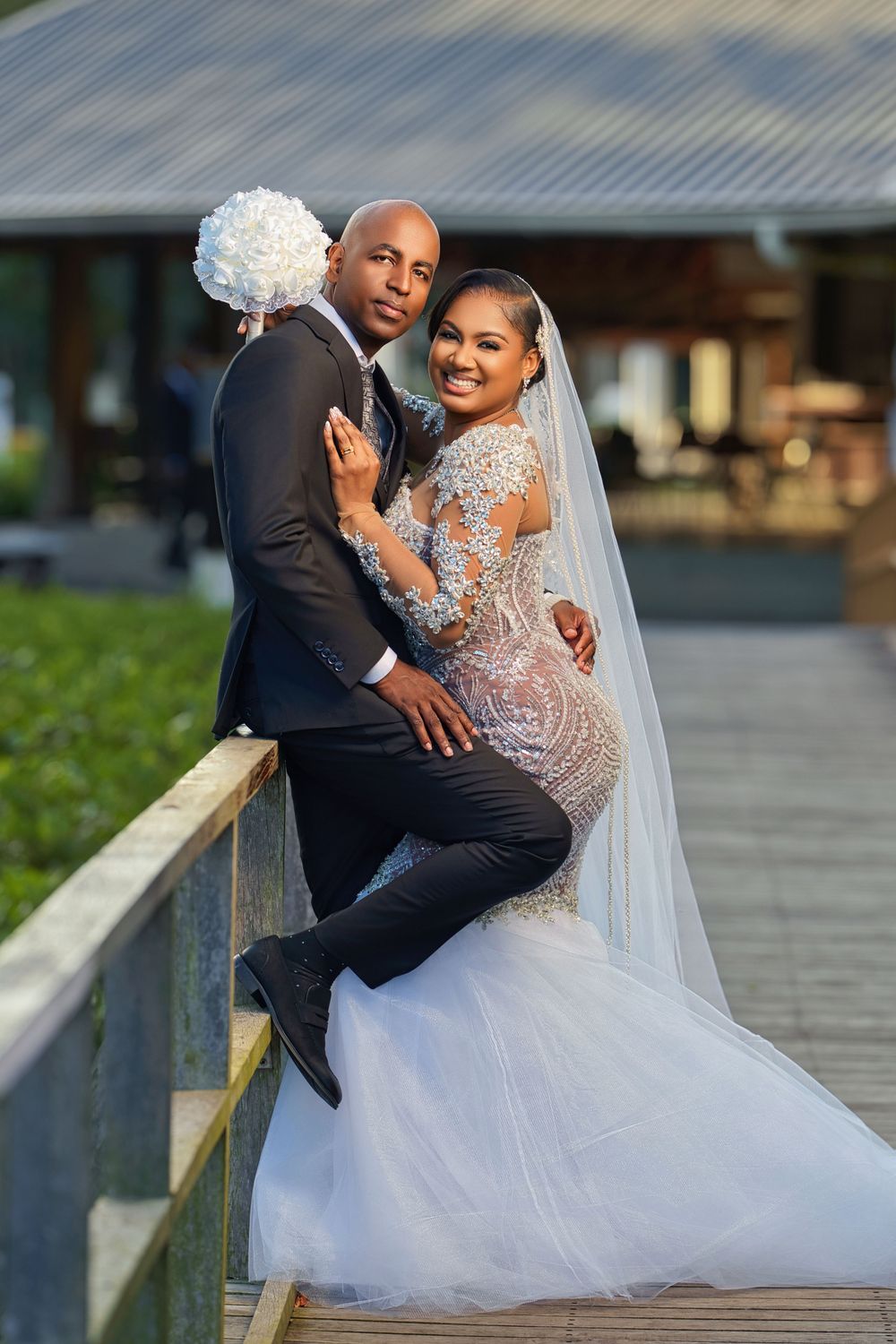 A bride in a beaded mermaid gown and veil poses with a groom in a black suit on a wooden deck.