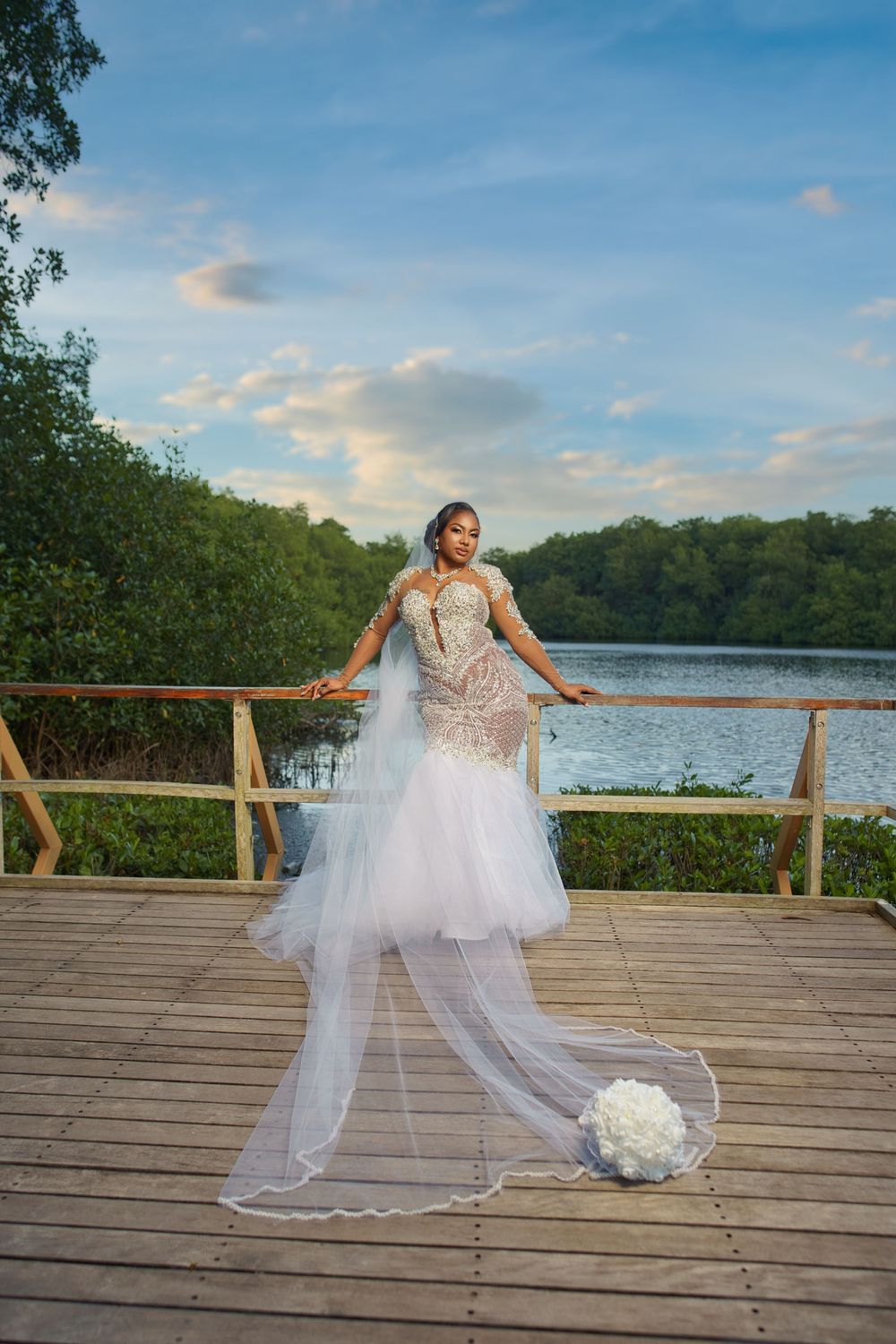 Bride in lace mermaid gown with veil poses on wooden deck overlooking serene lake surrounded by lush greenery.