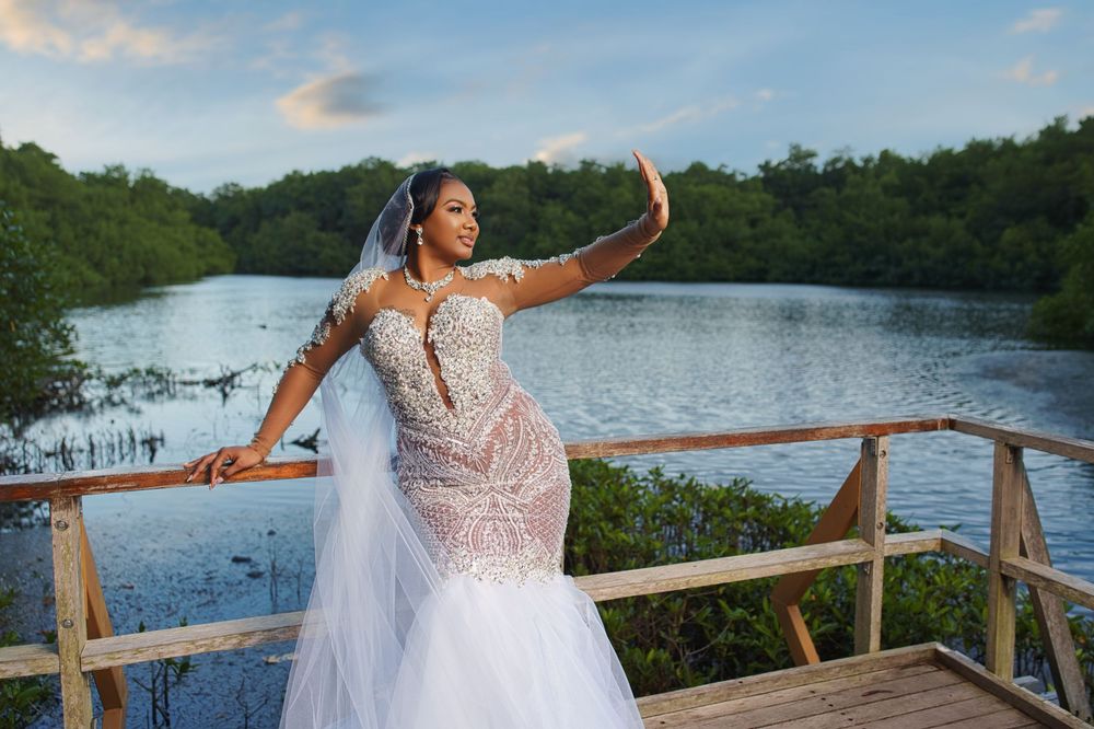 Bride in beaded mermaid gown and veil poses on wooden deck overlooking serene lake surrounded by lush greenery.