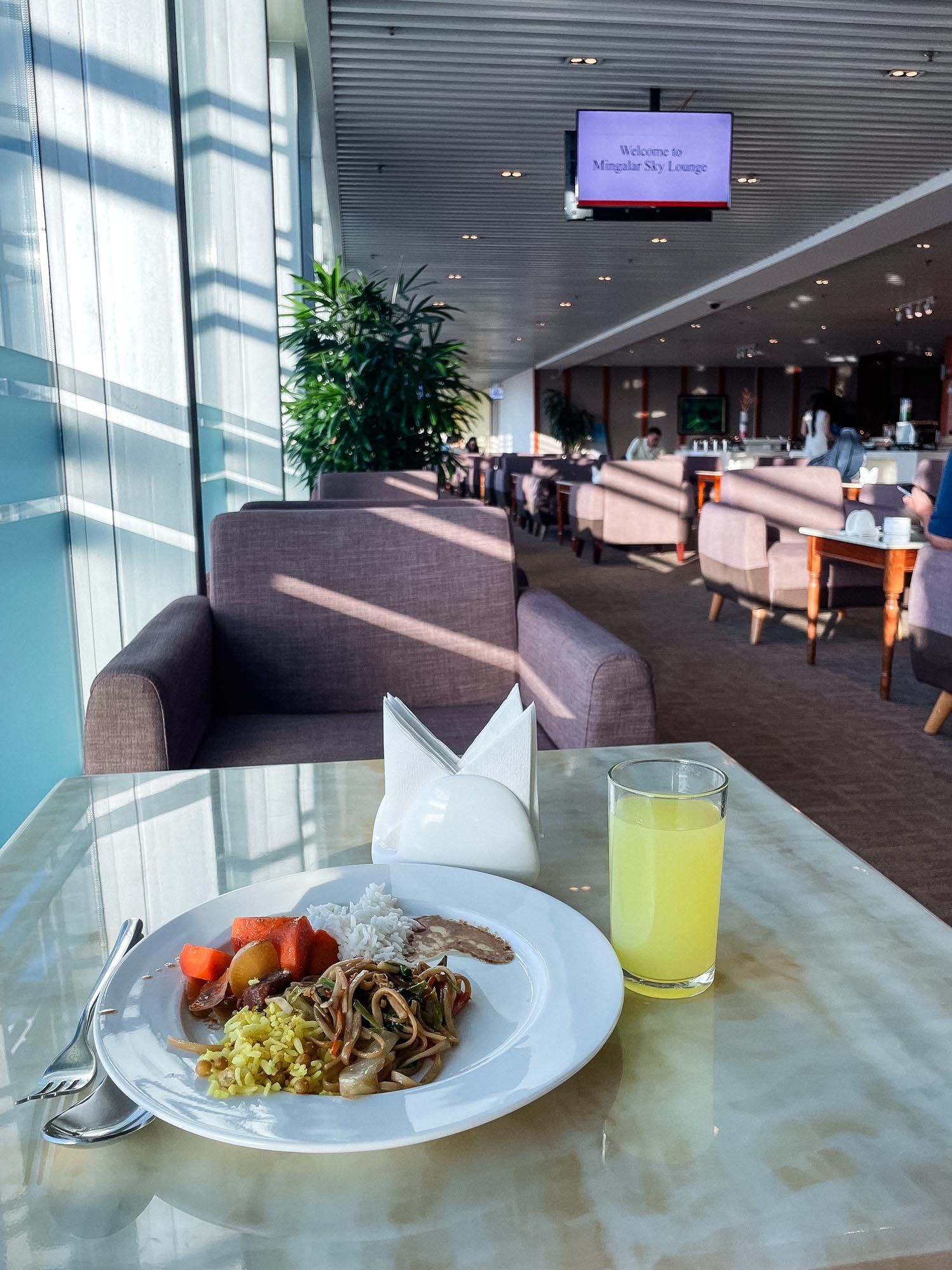 Airport lounge dining area with plated meal, yellow drink, and comfortable seating overlooking large windows.
