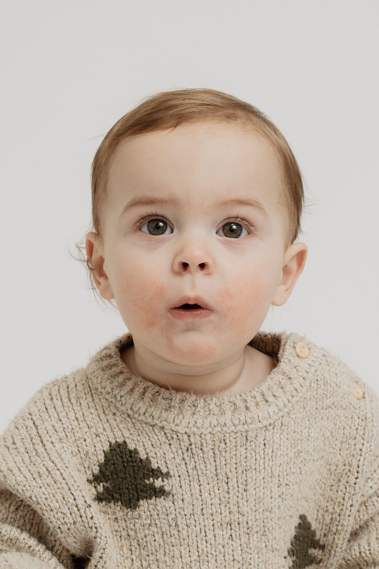 Baby wearing a cream knit sweater with dark pattern looks upward with curious expression.
