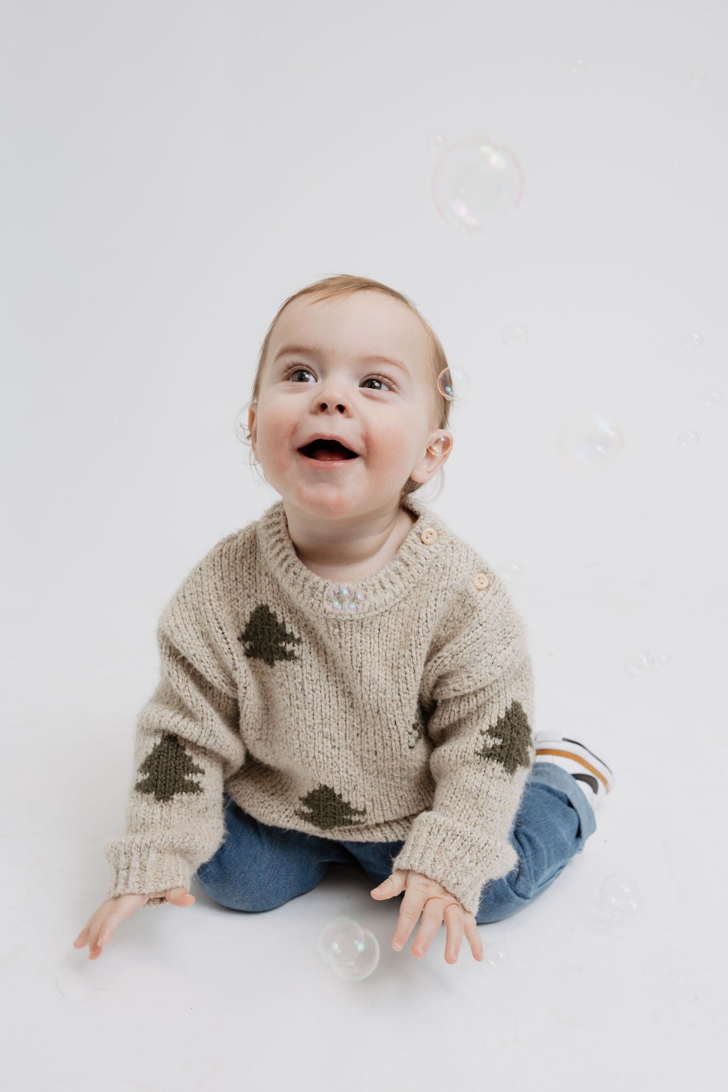 Baby wearing knit sweater with tree pattern and jeans sits on white background while looking at floating bubbles.