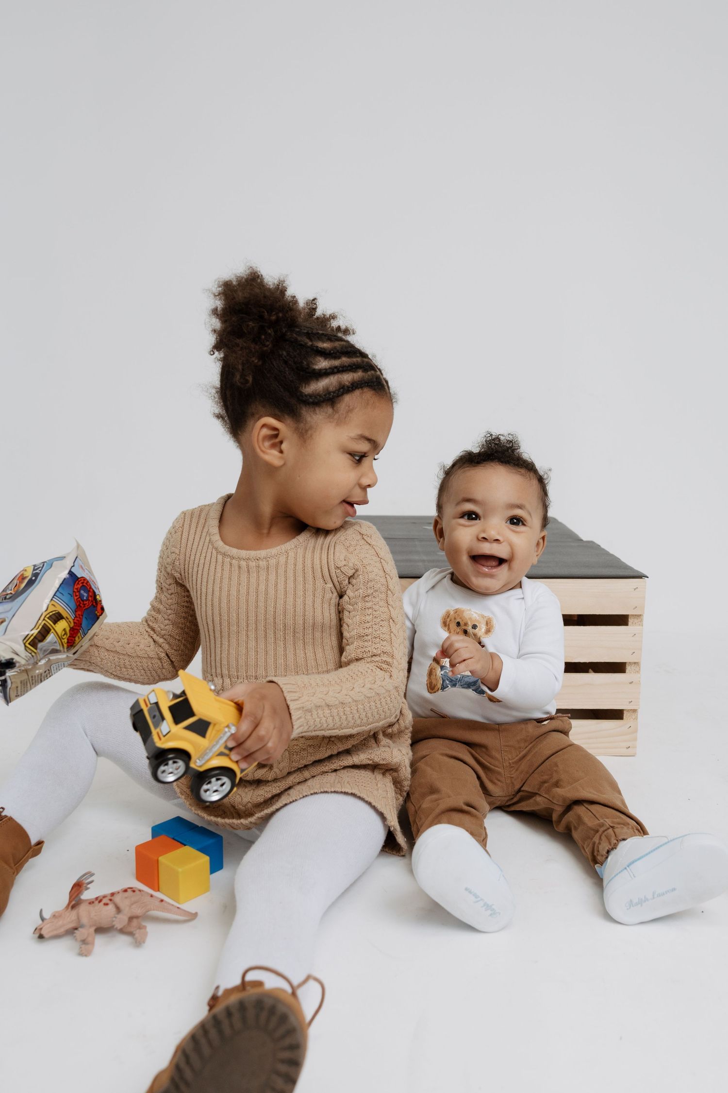Two children in matching beige sweaters play with colorful toy cars on a white surface while sharing joyful moments.