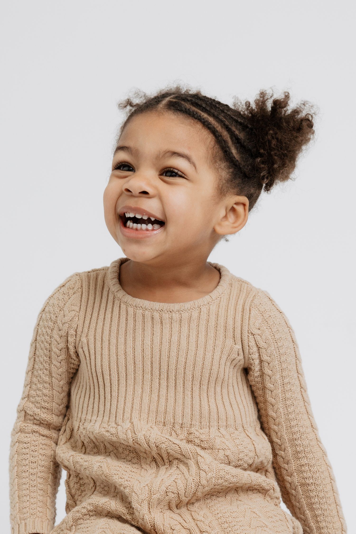 Young child wearing beige cable knit sweater smiles joyfully against white background.
