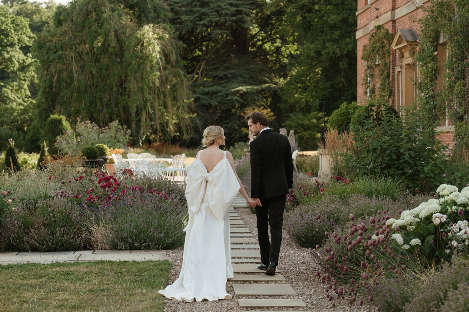 Bride and groom walk through garden path with red brick building in background.