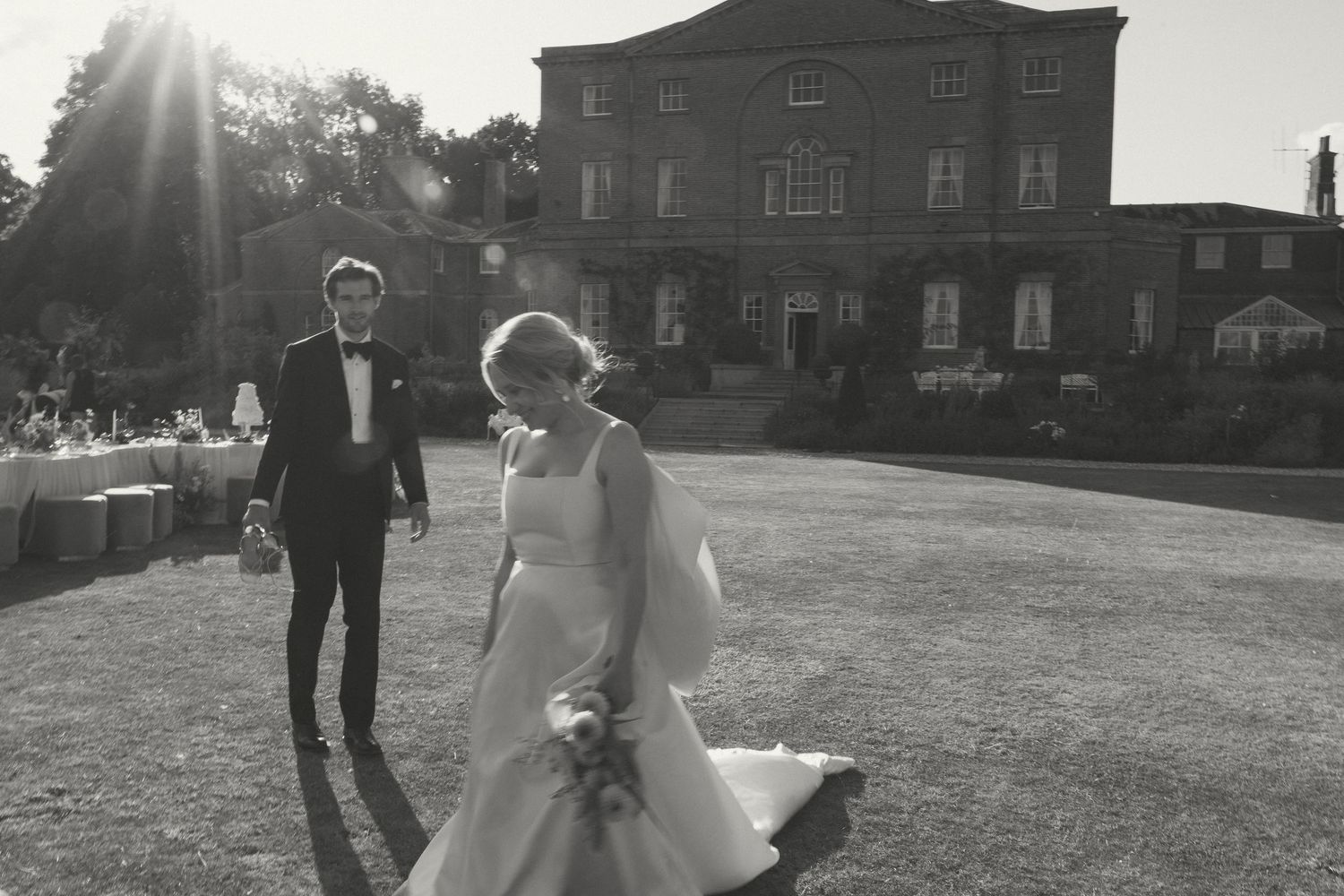 Black and white wedding photo of a couple walking on a lawn in front of a brick mansion with sunlight peeking through.