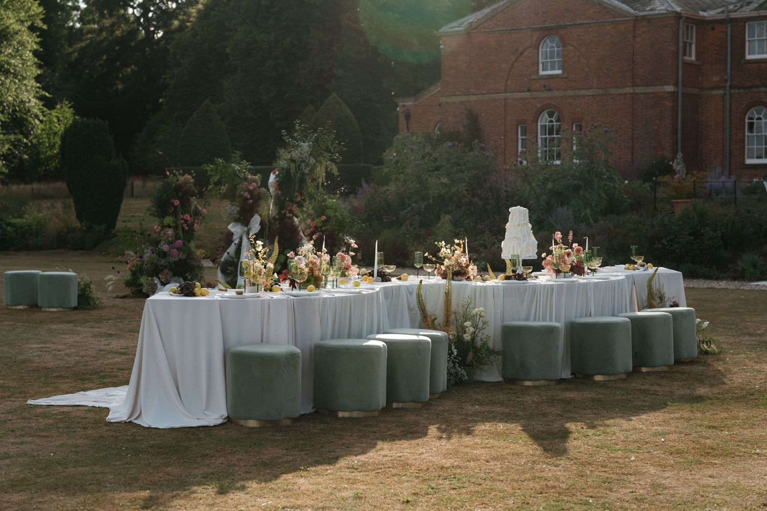 Elegant outdoor wedding reception setup with white tablecloths and sage green chairs on a lawn near a brick building.