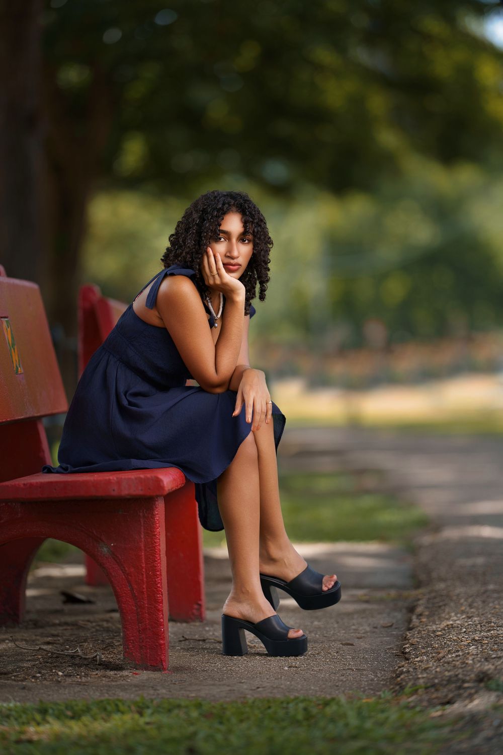 A figure in a navy dress sits thoughtfully on a red bench in a sunlit outdoor setting.