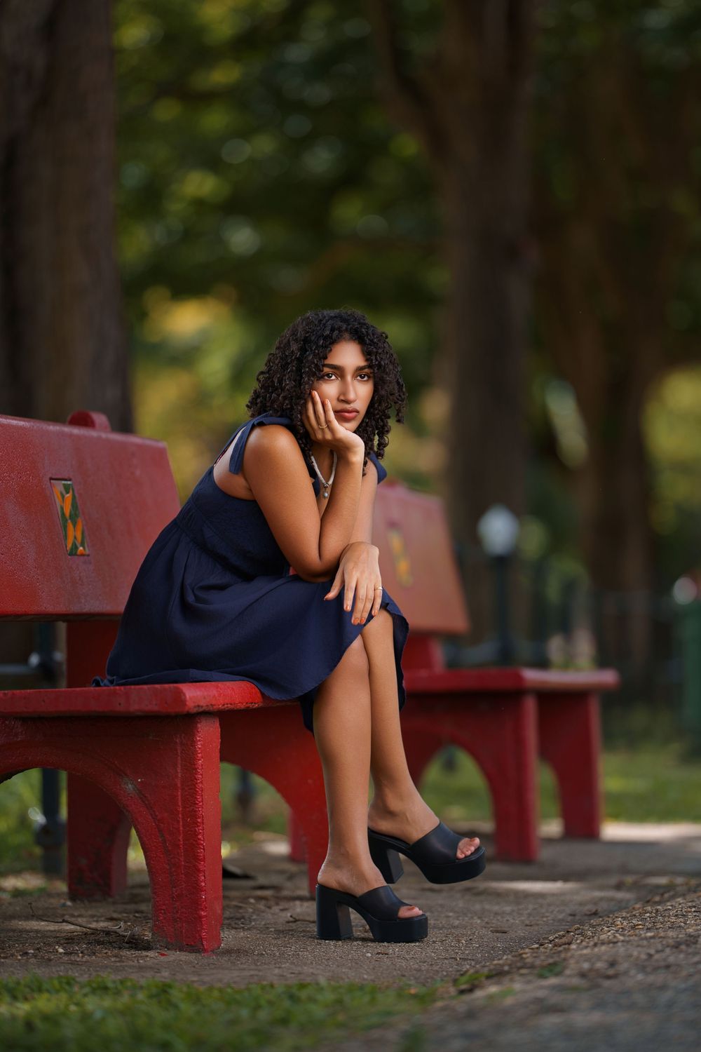 A person in a navy blue dress sits thoughtfully on a red park bench surrounded by trees in soft natural lighting.