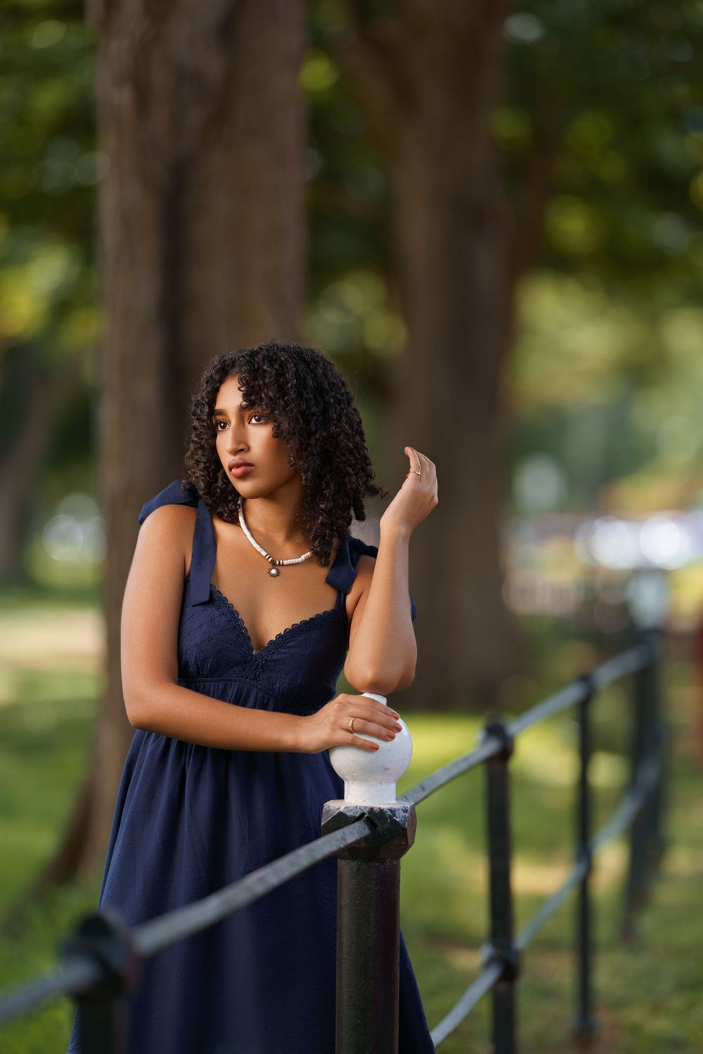 A woman in a navy blue dress poses thoughtfully against a railing in a park setting with soft natural lighting.
