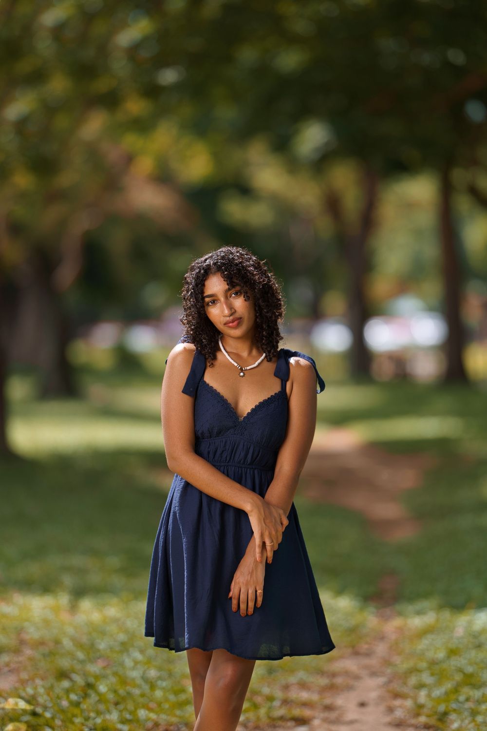 Woman in navy blue summer dress poses outdoors in a park with blurred green trees in the background.