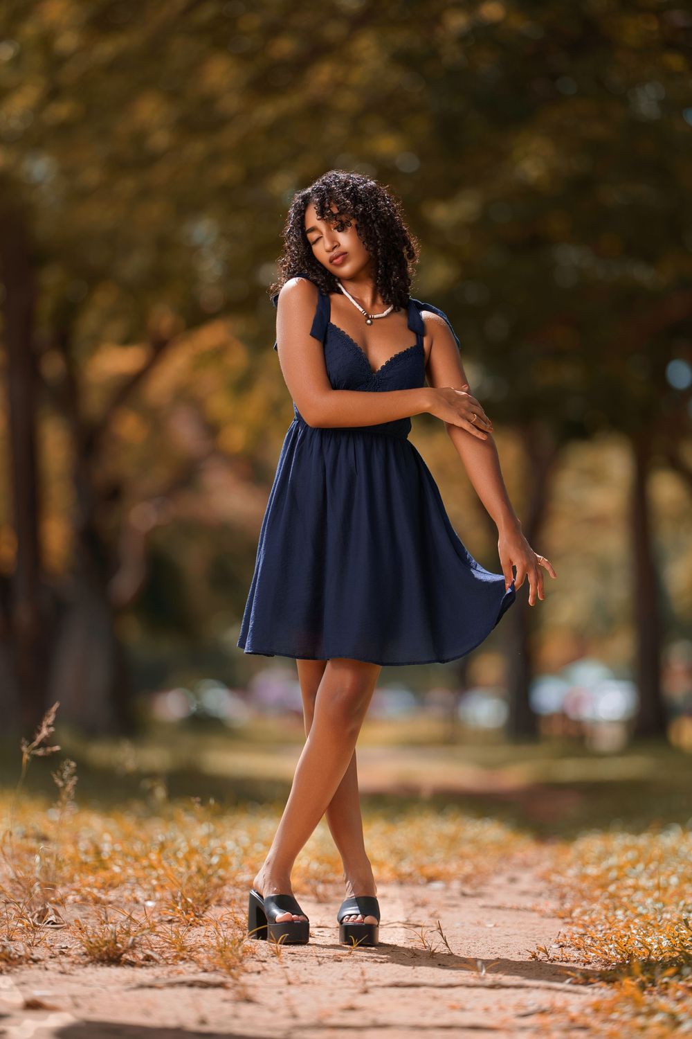 A person in a navy blue dress stands playfully on a path covered in fallen autumn leaves.