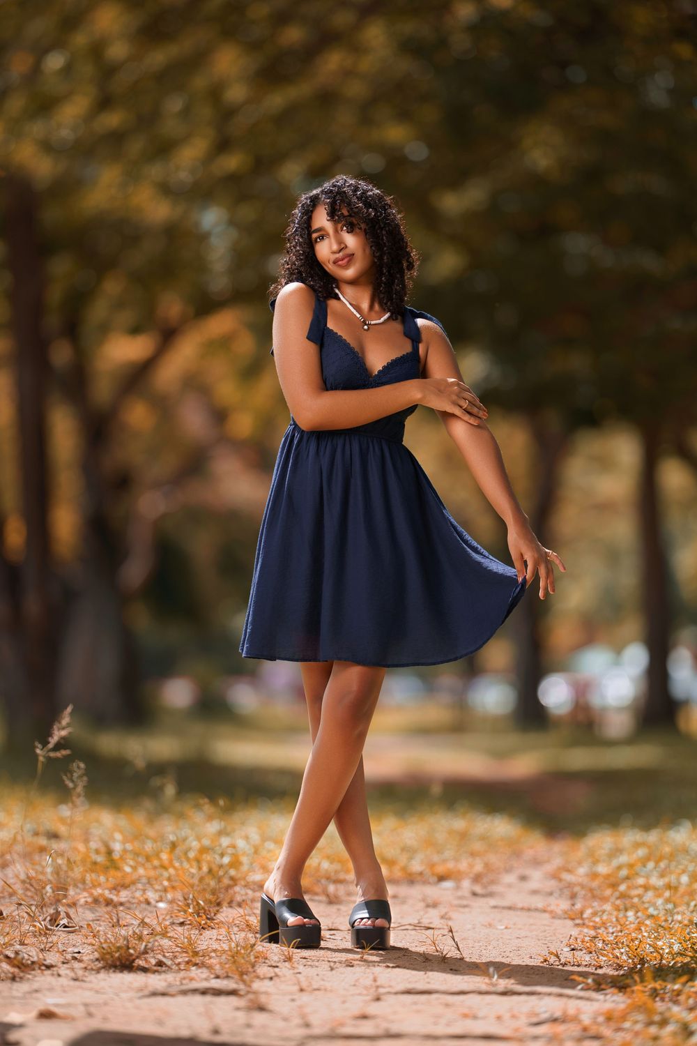 A person in a navy blue dress poses playfully among autumn leaves on a tree-lined path.