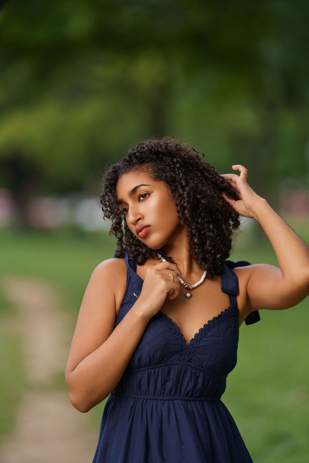 A woman in a dark dress stands elegantly in an outdoor setting with lush greenery and trees in the background.