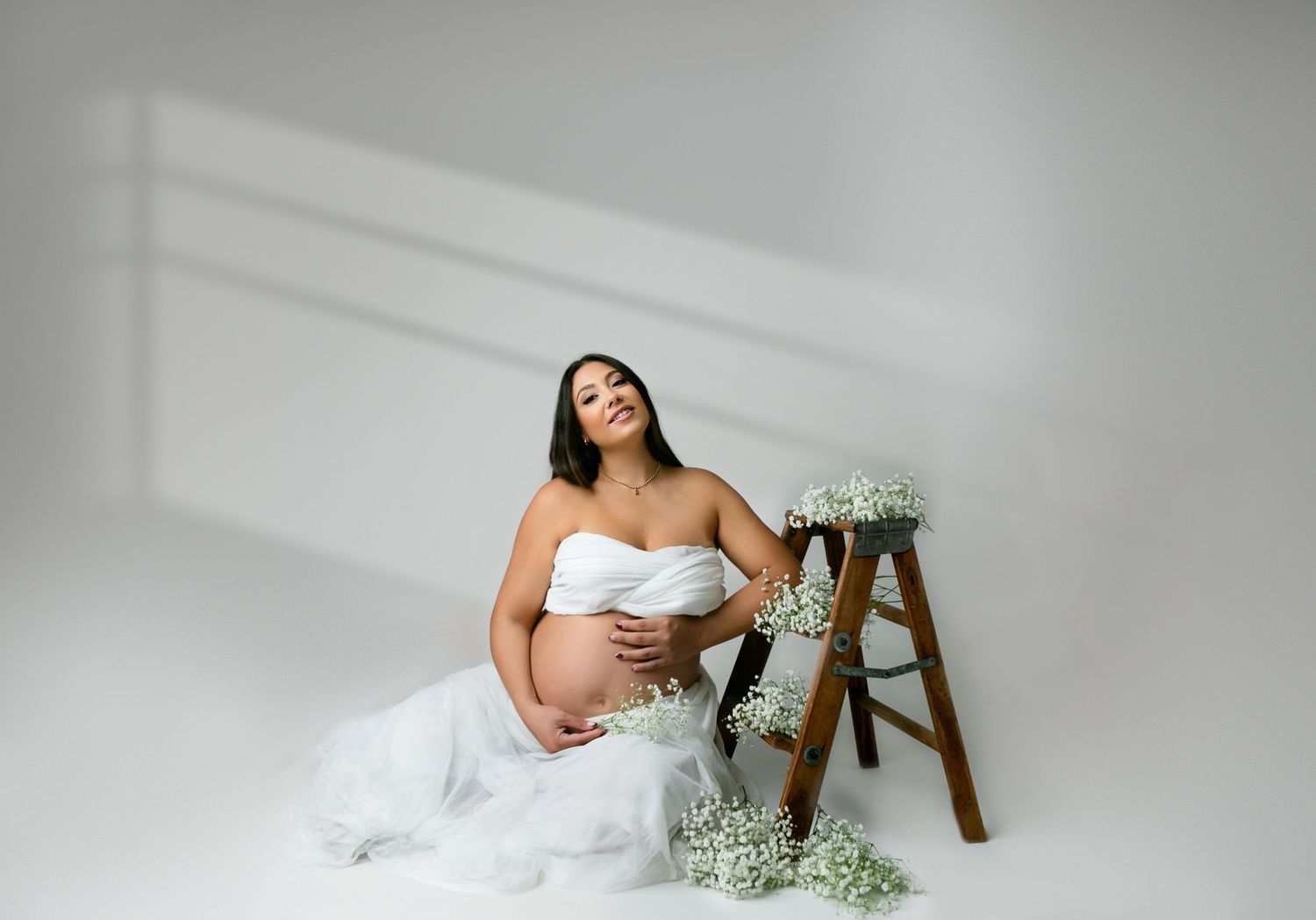 A maternity session showing a mom in a white dress with baby's breath flowers against a light backdrop. Orlando Maternity Photography