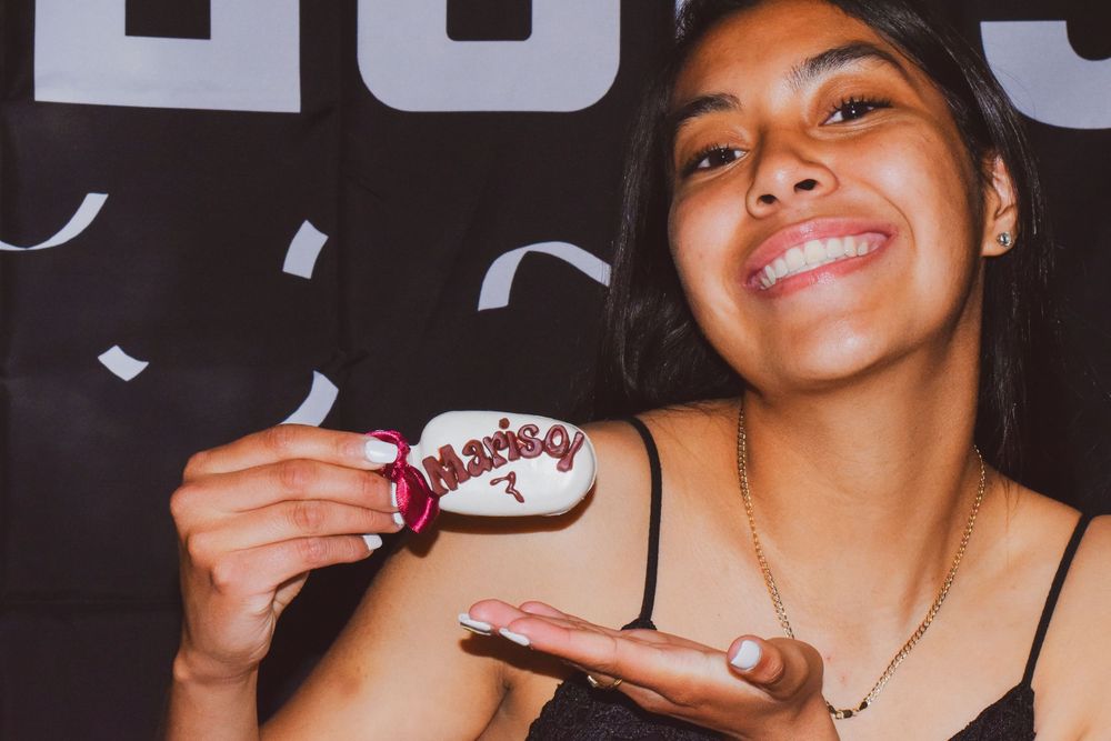 A person smiles while holding a small cupcake with white frosting and colorful sprinkles.