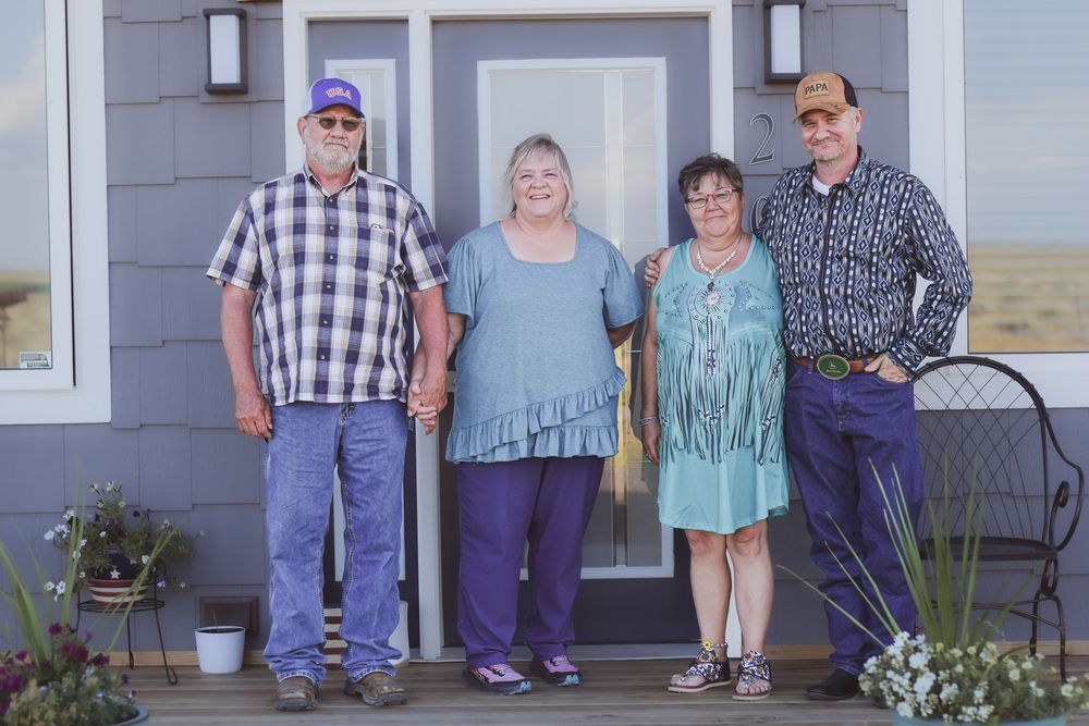 People stand together on a porch of a house with white siding and decorative plants outside.