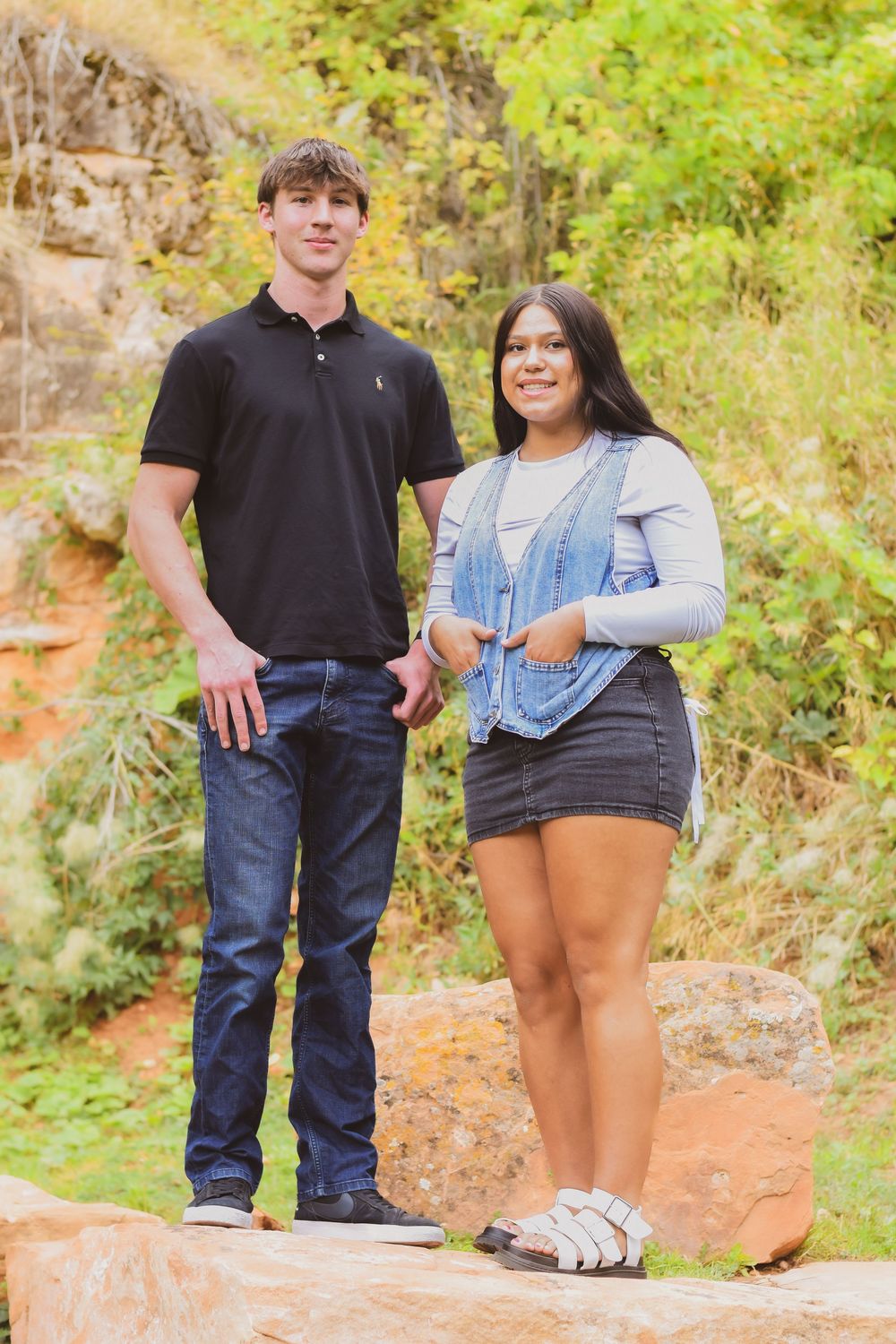 A couple stands together on a red clay path surrounded by lush green foliage in a natural outdoor setting.