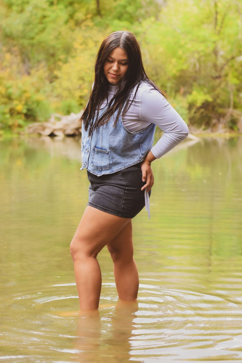 A person in denim shorts and gray top wades into a tranquil lake surrounded by lush green trees.