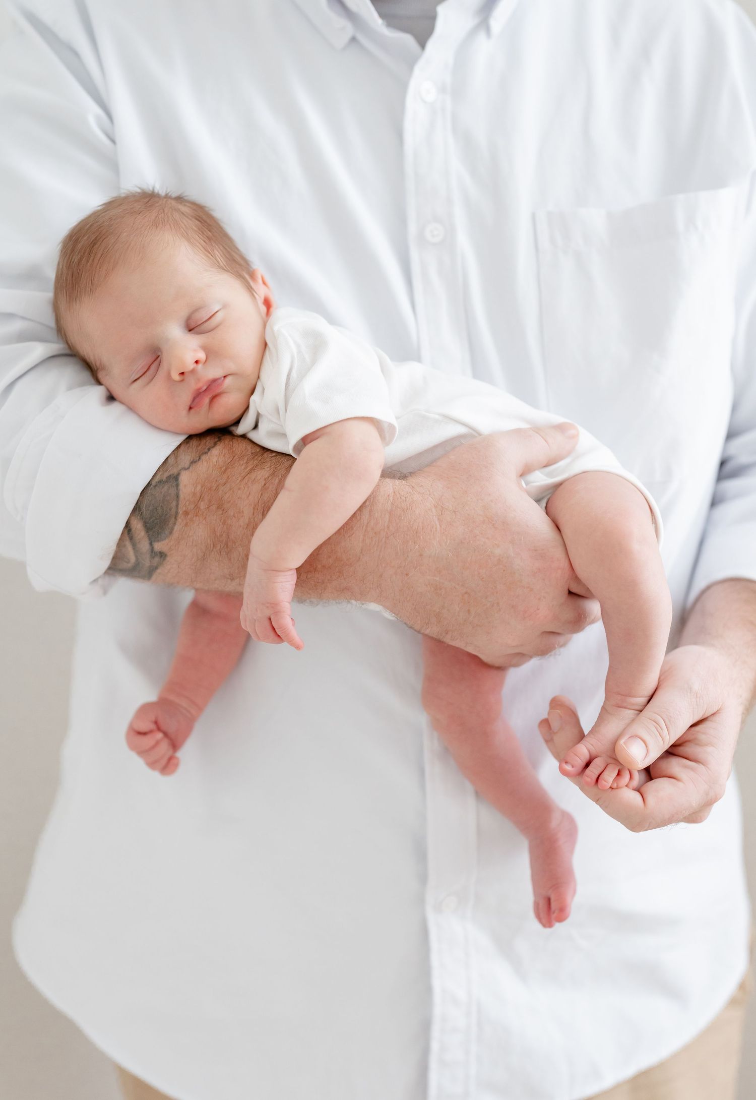 A sleeping newborn baby lying on white bedding while being gently held by adult hands.