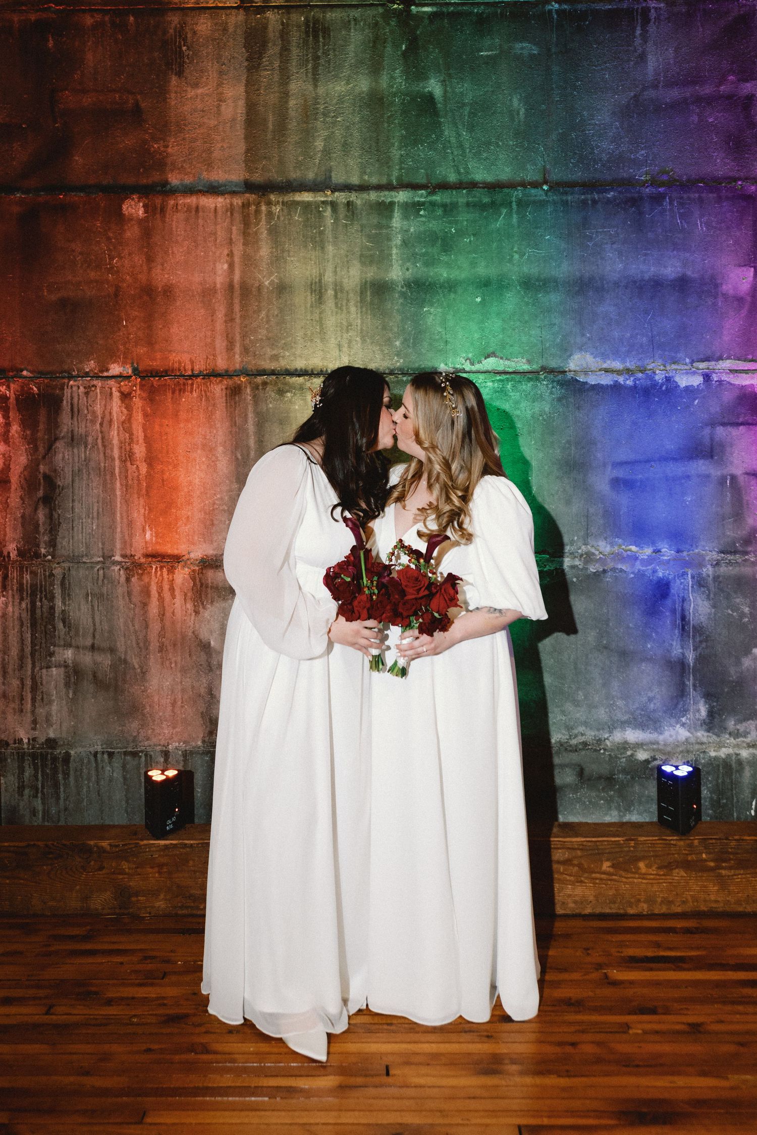 A rainbow-lit backdrop illuminates two people in white dresses holding red bouquets during their special moment.