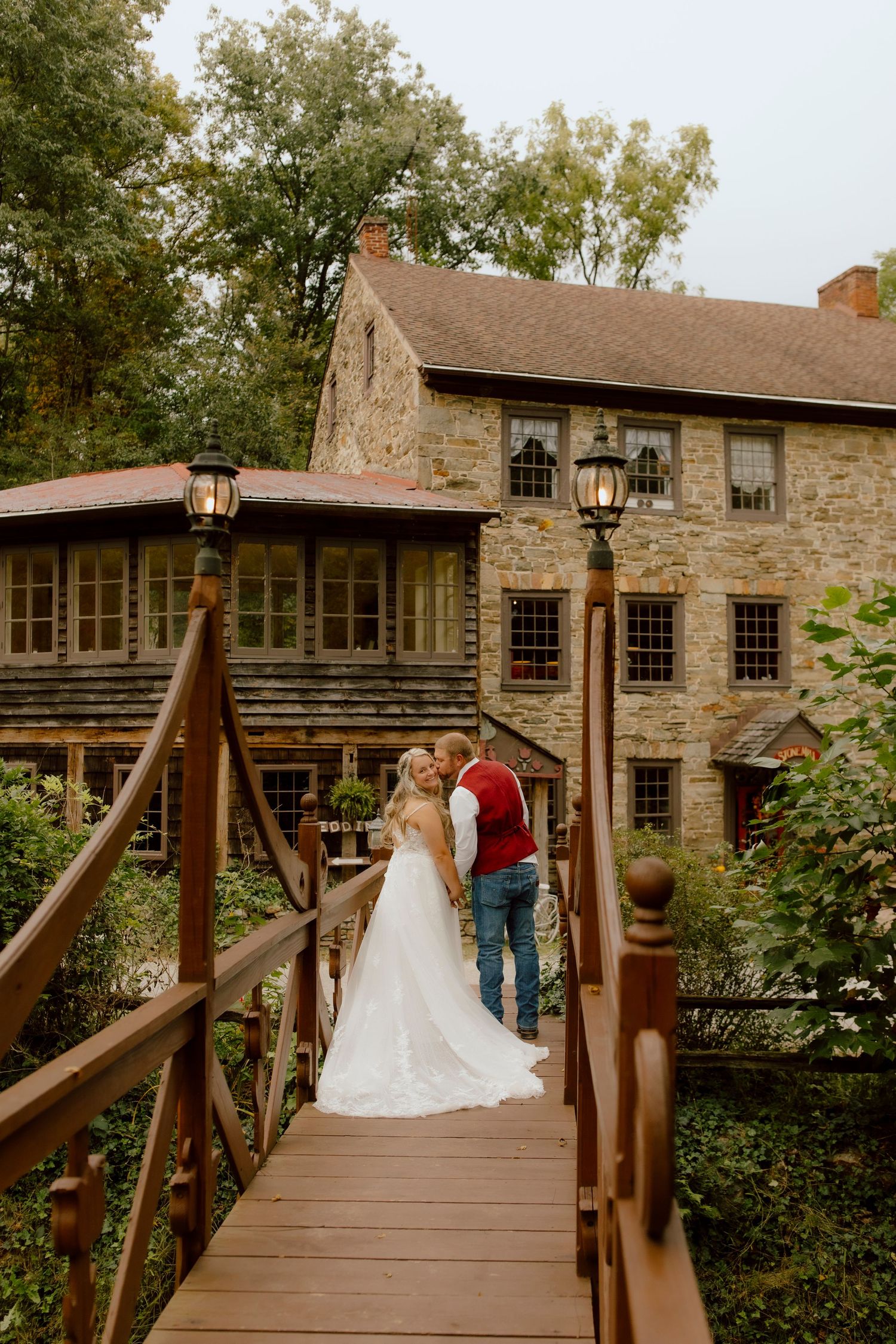 Bride and groom share a kiss on a wooden bridge outside a rustic stone building.