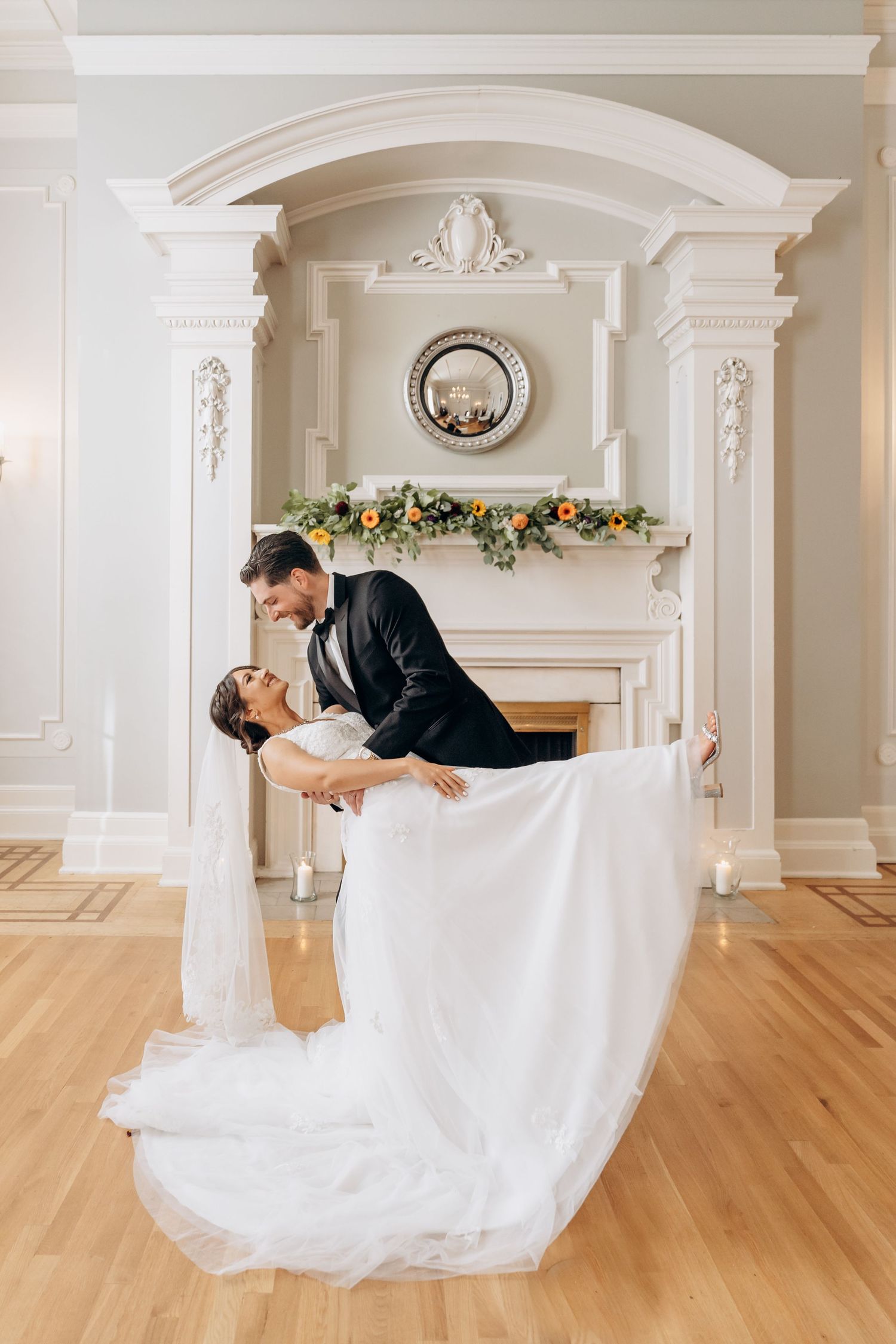 A wedding couple performs a dip dance sequence in an elegant white ballroom with ornate architectural details.