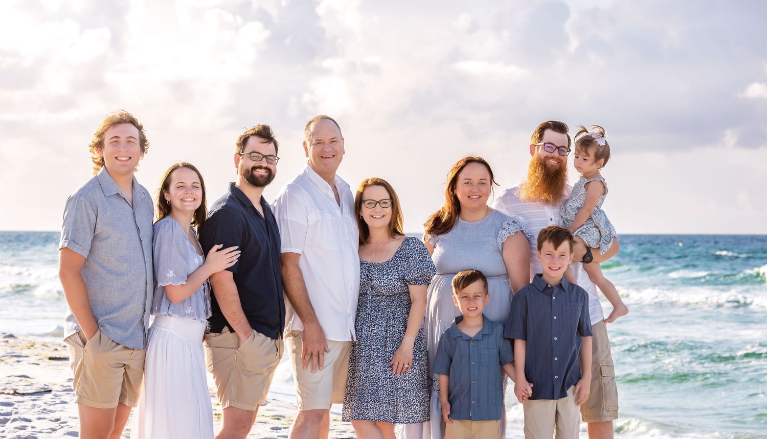 A group of people wearing coordinated blue and white outfits pose together on a sunny beach for family photos.