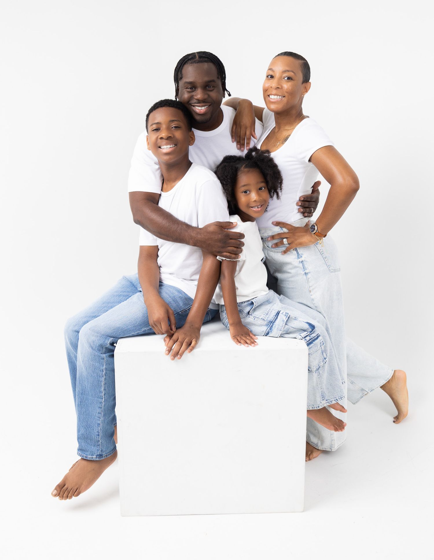 A family wearing white shirts and blue jeans poses together on a white cube against a light background.