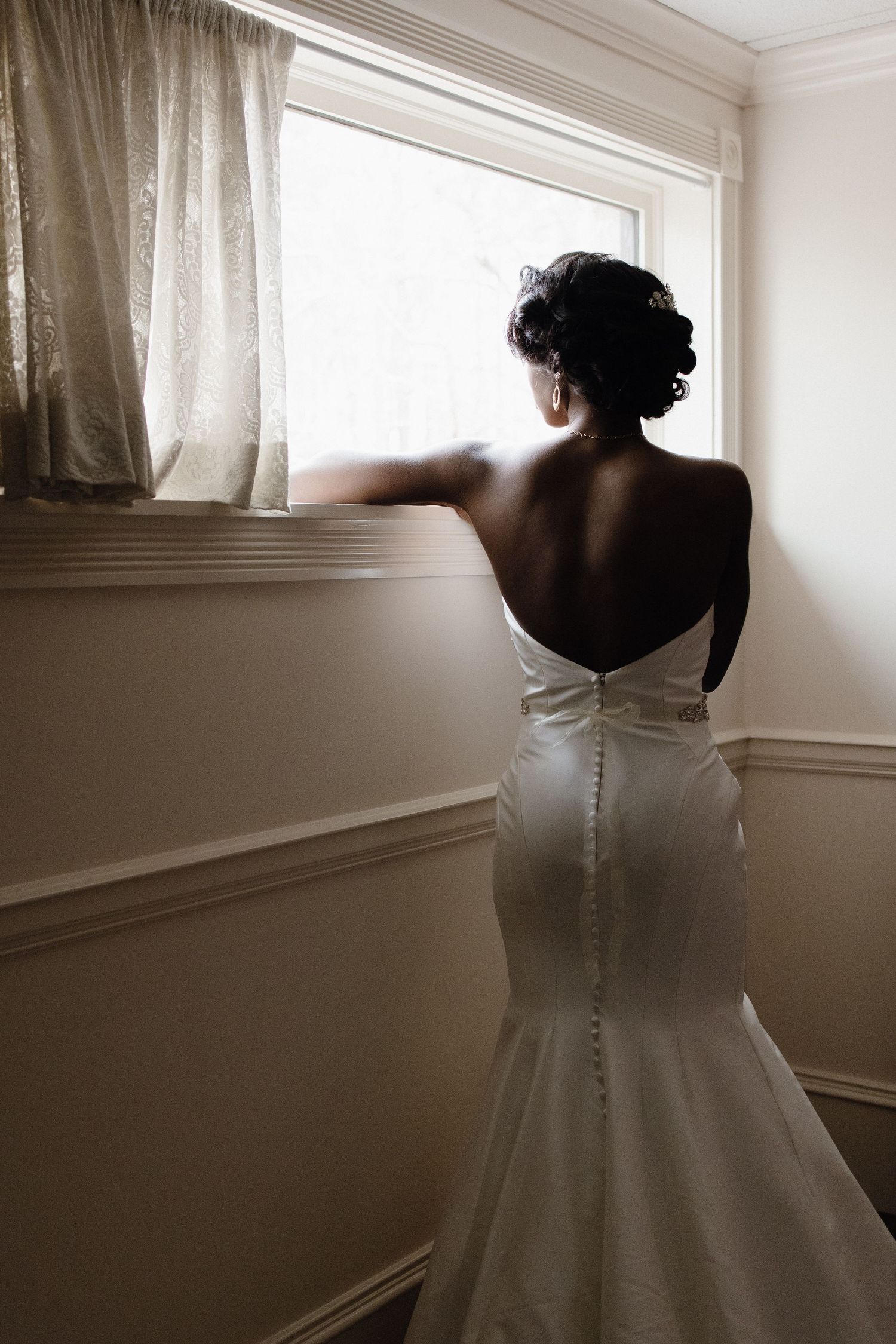 Silhouetted view of a wedding dress from behind in a sunlit room with white curtains.